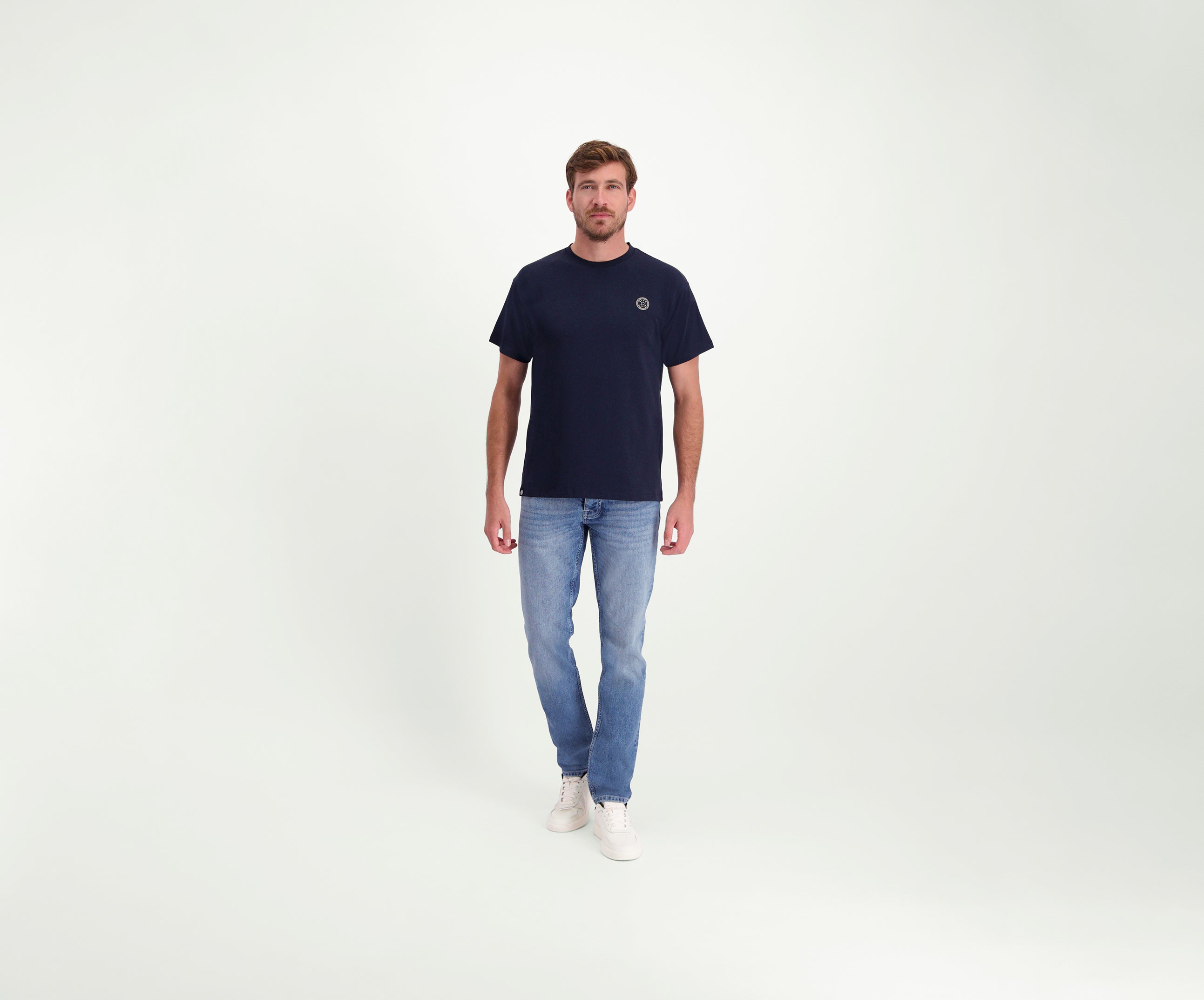 Een man draagt The N01 Tee Navy van Heren, lichtblauwe Blue Rivet jeans en regular-fit witte sneakers en staat tegen een effen witte achtergrond. Hij kijkt recht in de camera met een neutrale uitdrukking.