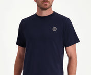 Een man draagt The N01 Tee Navy van Blue Rivet Jeans, een regular fit t-shirt in effen marineblauw met een klein rond embleem op de linkerborst. Hij poseert met zijn armen langs zijn lichaam tegen een effen witte achtergrond.