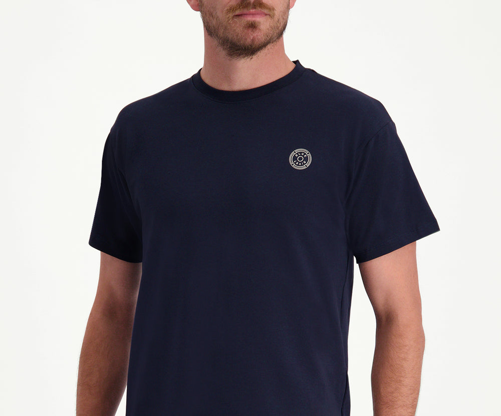Een man met kort haar, gekleed in de kleuren The N01 Tee Navy en Blue Rivet Jeans, staat met zijn handen op zijn rug tegen een effen witte achtergrond.