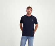 Een man met kort haar, gekleed in de kleuren The N01 Tee Navy en Blue Rivet Jeans, staat met zijn handen op zijn rug tegen een effen witte achtergrond.