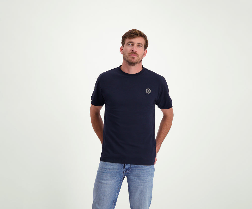 Een man met kort haar, gekleed in de kleuren The N01 Tee Navy en Blue Rivet Jeans, staat met zijn handen op zijn rug tegen een effen witte achtergrond.