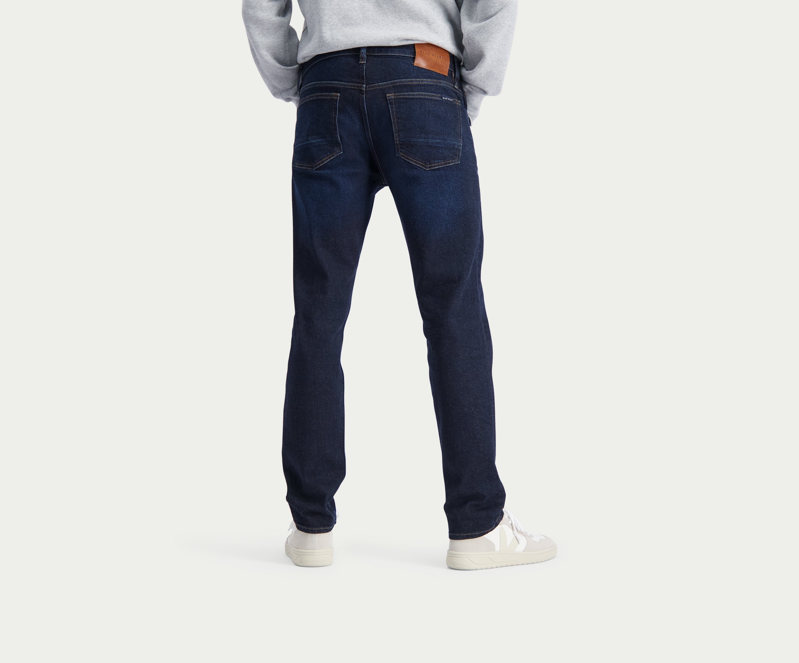 Een persoon draagt de N01 Slim Dark Indigo jeans van Blue Rivet Jeans en witte sneakers en staat met zijn rug naar de camera tegen een effen witte achtergrond.