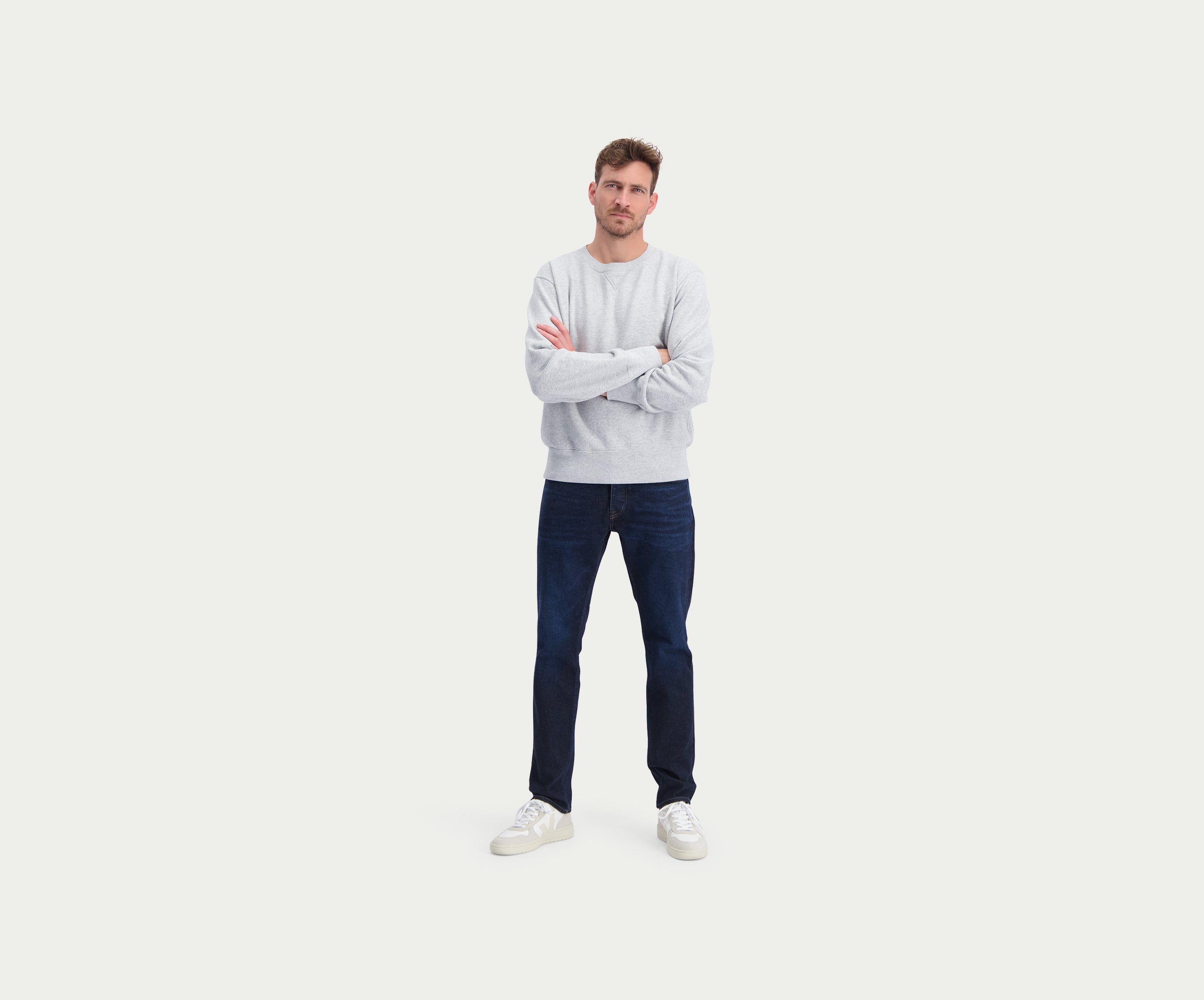 Een man staat zelfverzekerd met gekruiste armen, gekleed in een grijze trui van topkwaliteit, "The N01 Slim Dark Indigo" van Blue Rivet Jeans en witte sneakers tegen een effen witte achtergrond.