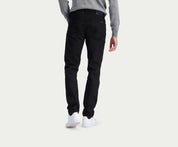 Een persoon die de N01 Slim Black jeans van Blue Rivet Jeans draagt, staat met zijn rug naar de camera. Hij draagt een grijs shirt met lange mouwen en witte sneakers tegen een effen, lichte achtergrond voor een strakke en premium look.