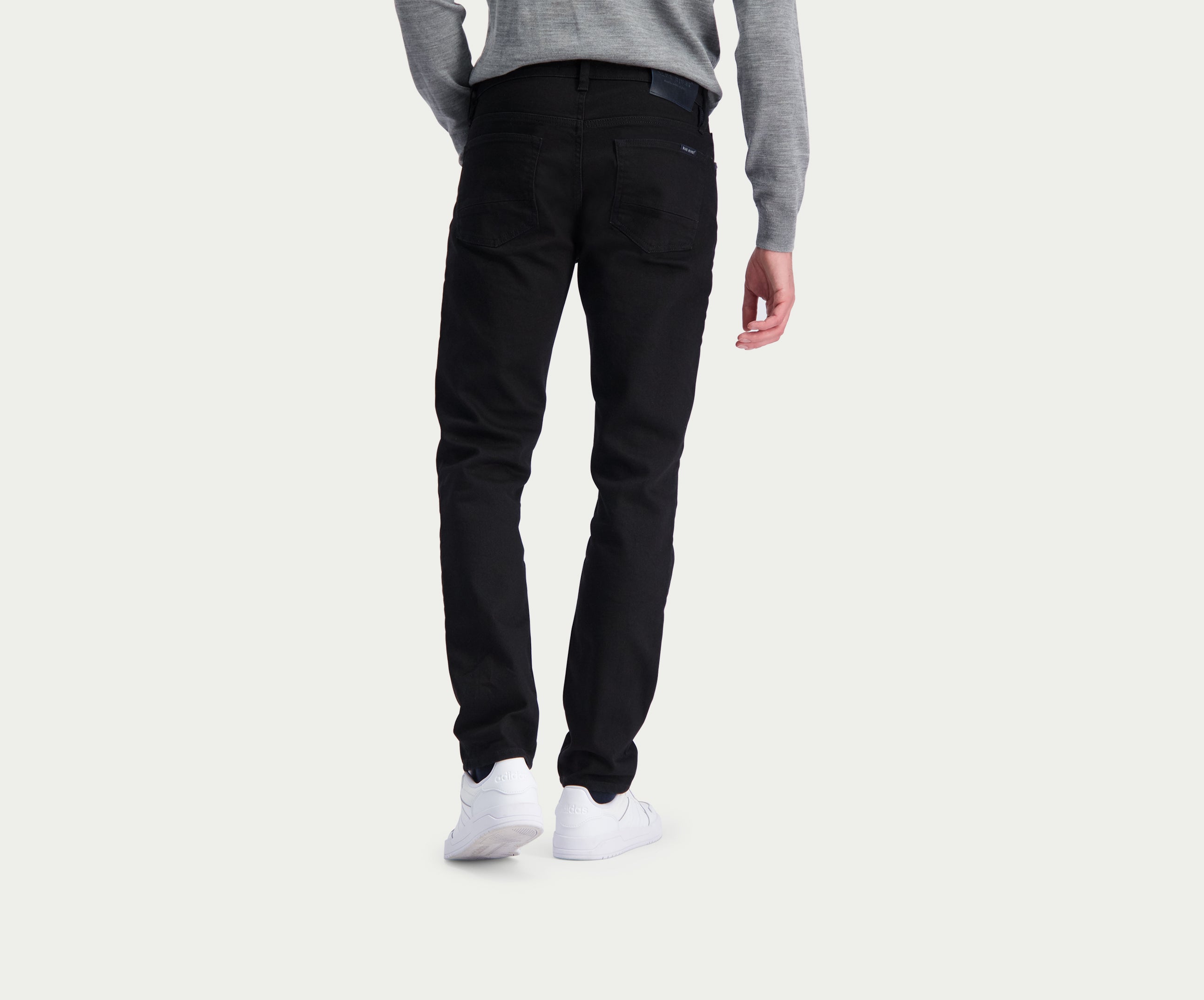 Een persoon die de N01 Slim Black jeans van Blue Rivet Jeans draagt, staat met zijn rug naar de camera. Hij draagt een grijs shirt met lange mouwen en witte sneakers tegen een effen, lichte achtergrond voor een strakke en premium look.