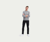 Een man staat met gekruiste armen, draagt een premium grijze trui, de N01 Slim Black broek van Blue Rivet Jeans en witte sneakers. Hij heeft kort haar en kijkt vooruit, tegen een effen witte achtergrond.