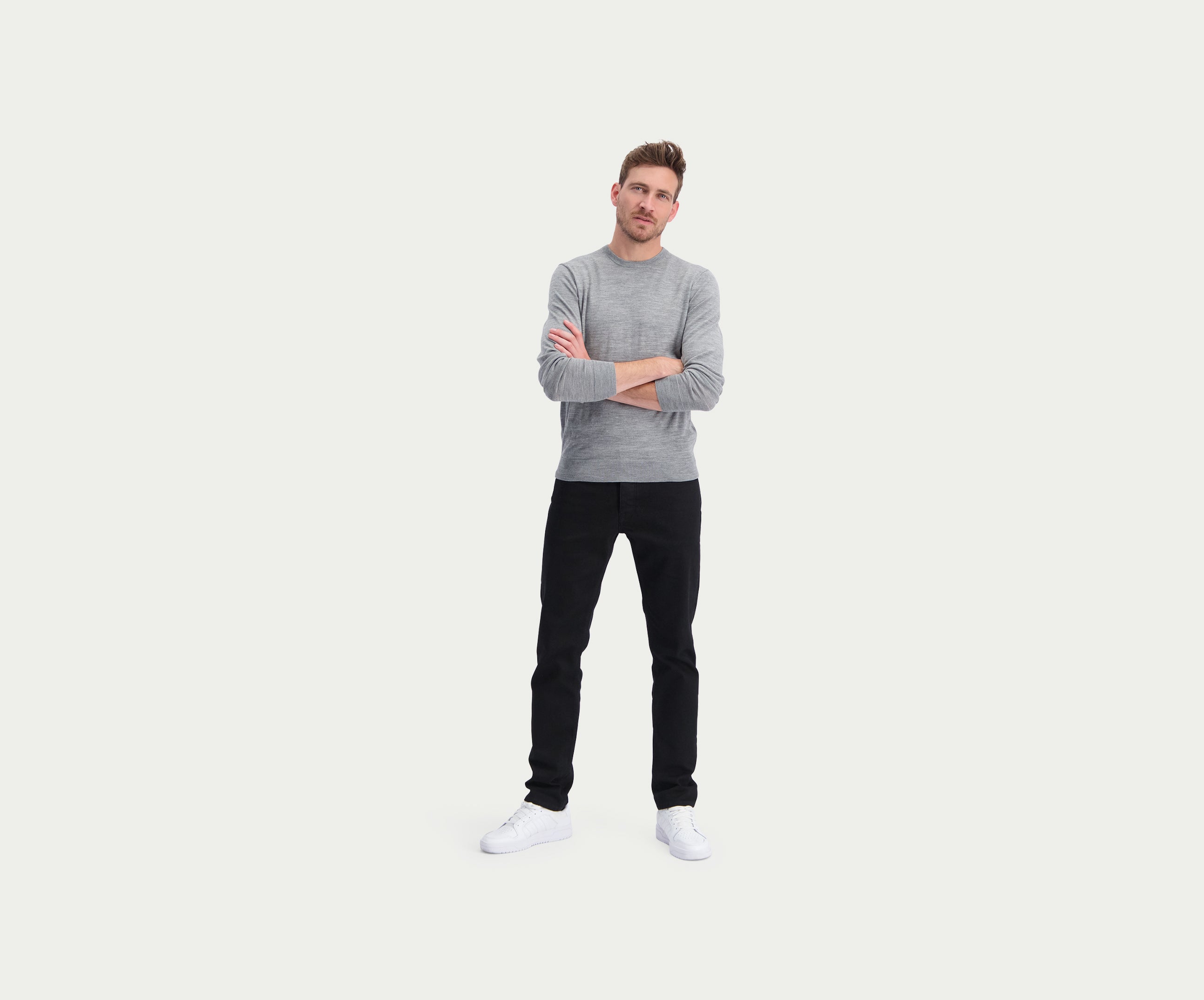 Een man staat met gekruiste armen, draagt een premium grijze trui, de N01 Slim Black broek van Blue Rivet Jeans en witte sneakers. Hij heeft kort haar en kijkt vooruit, tegen een effen witte achtergrond.