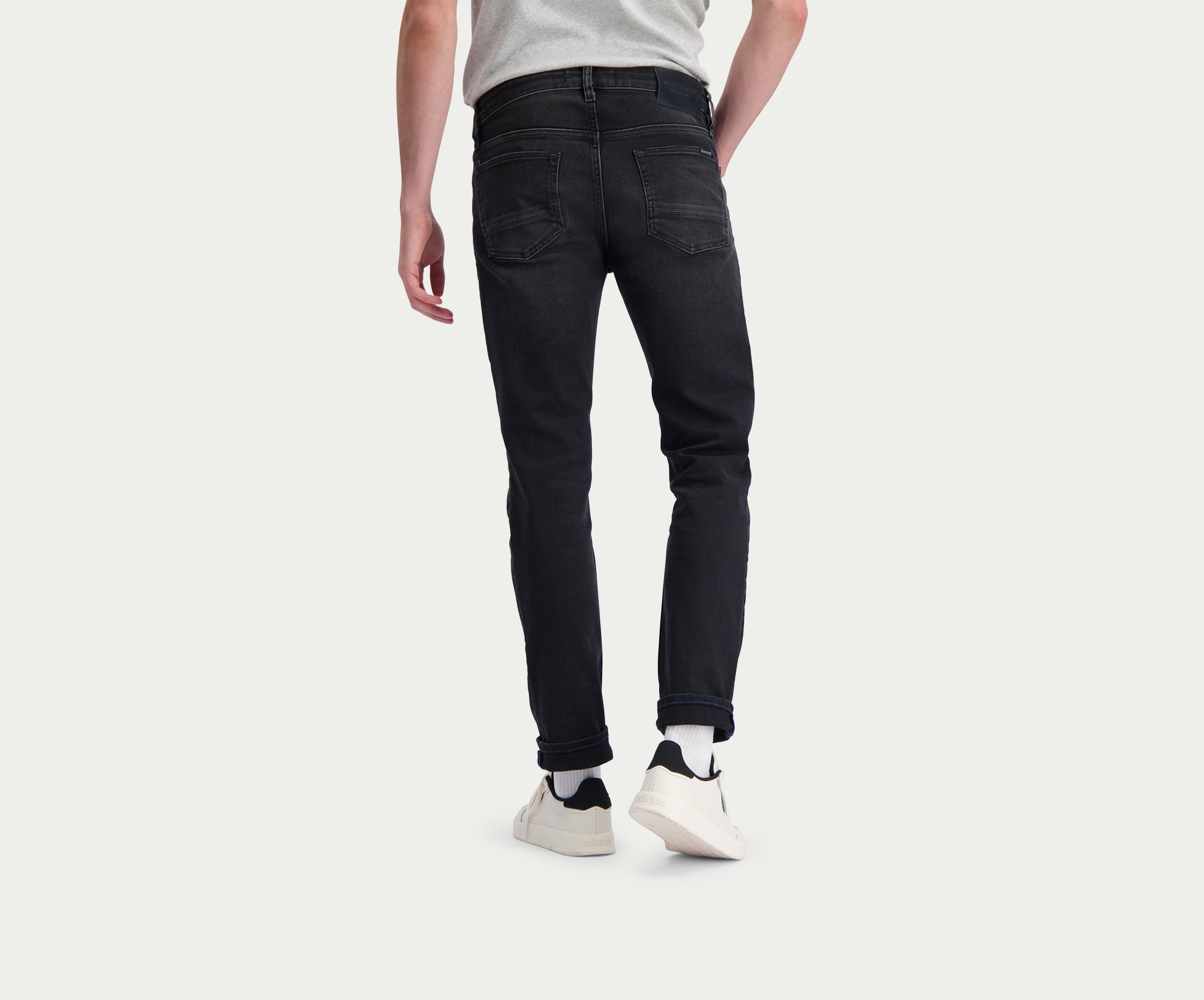 Een persoon die een lichtgrijs shirt draagt en Blue Rivet Jeans' The N01 Slim Black Used staat met zijn rug naar de camera. Hij draagt witte sneakers met zwarte accenten, tegen een effen witte achtergrond.
