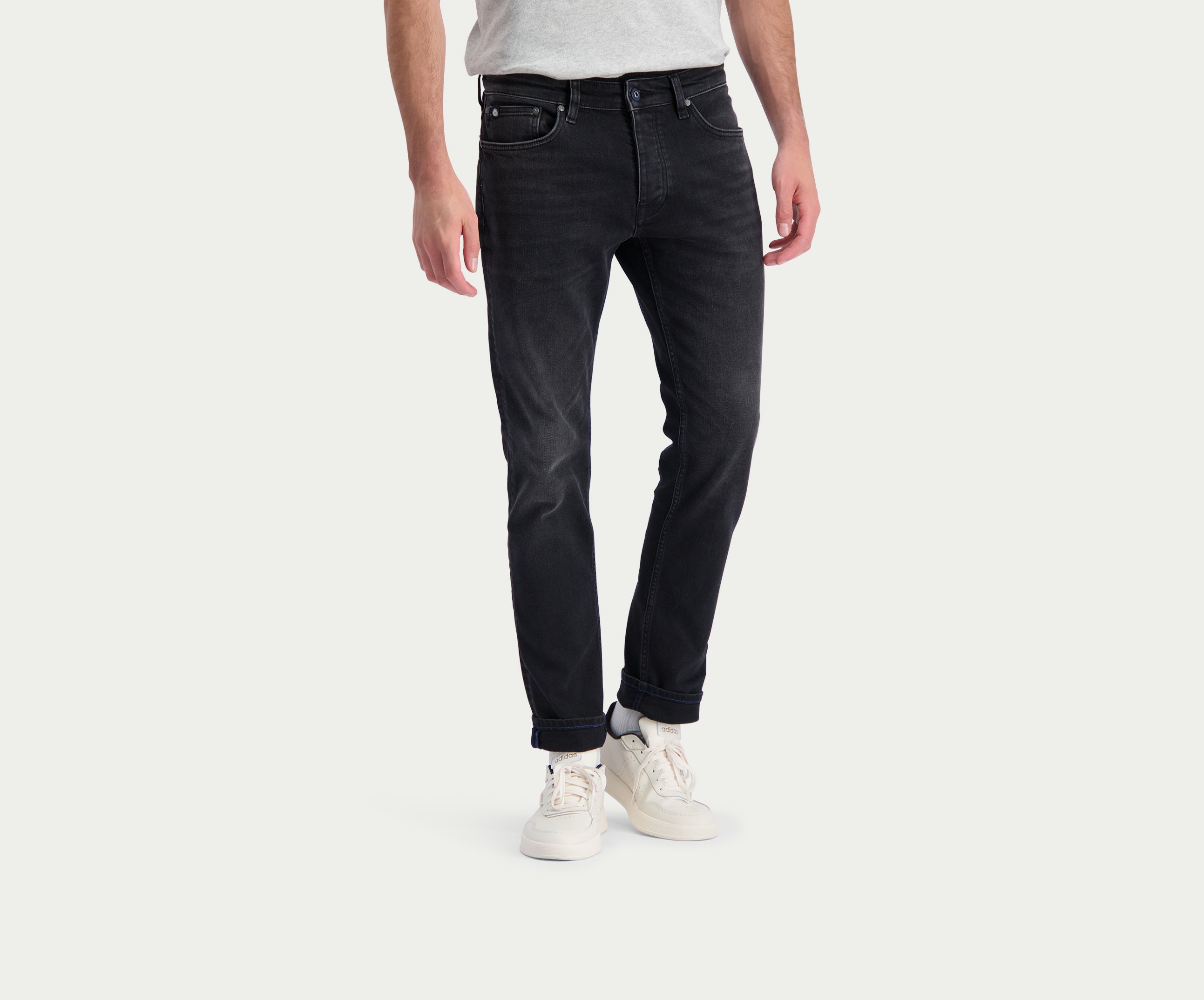 Een persoon die The N01 Slim Black Used jeans van Blue Rivet Jeans en witte sneakers draagt, staat tegen een effen lichte achtergrond. De zwarte jeans worden gecombineerd met een grijs t-shirt en de handen van de persoon liggen ontspannen langs hun zijden.
