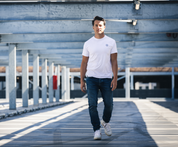 Iemand draagt de witte The N01 Tee van Blue Rivet Jeans en een normale blauwe spijkerbroek en loopt op een zonnige, moderne parkeerplaats, waar metalen balken en schaduwen een geometrisch patroon op de grond creëren.