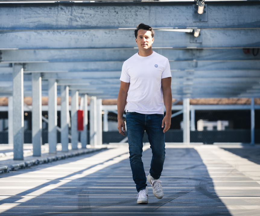 Iemand draagt de witte The N01 Tee van Blue Rivet Jeans en een normale blauwe spijkerbroek en loopt op een zonnige, moderne parkeerplaats, waar metalen balken en schaduwen een geometrisch patroon op de grond creëren.