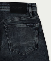 Close-up van The N01 Slim Blue Black Used denim jeans van Blue Rivet Jeans, met een zichtbare achterzak met een klein label erboven. De slanke tailleband is versierd met een leren patch met de merknaam Blue Rivet Jeans in reliëf. Deze jeans hebben een klassieke, vervaagde uitstraling.
