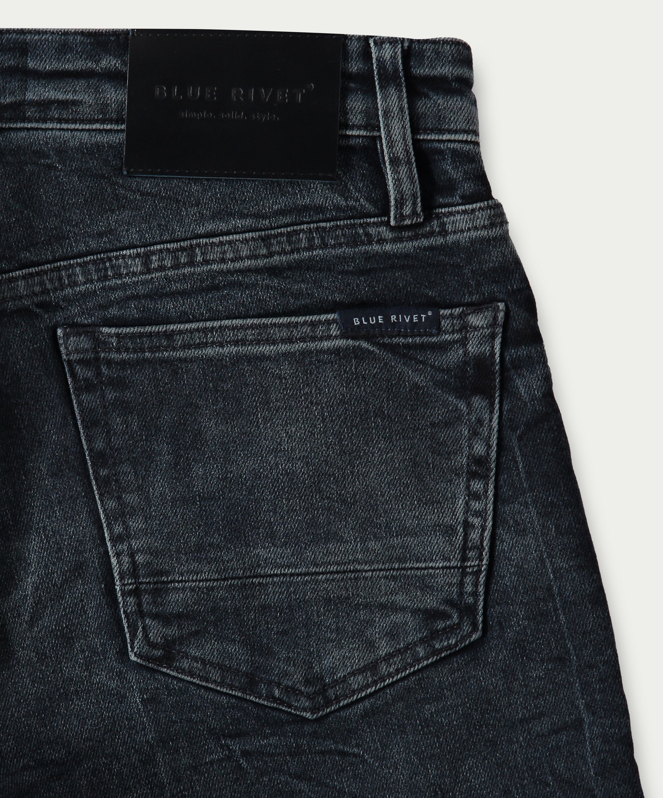 Close-up van The N01 Slim Blue Black Used denim jeans van Blue Rivet Jeans, met een zichtbare achterzak met een klein label erboven. De slanke tailleband is versierd met een leren patch met de merknaam Blue Rivet Jeans in reliëf. Deze jeans hebben een klassieke, vervaagde uitstraling.