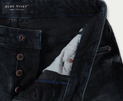 Close-up van donkerblauwe slim jeans met een zichtbare knoopsluiting. Het label luidt Blue Rivet Jeans met de tagline simple. solid. style. Koperen klinknagelknopen zijn prominent aanwezig en de binnenstof heeft een contrasterende lichtere kleur. De N01 Slim Blue Black Used spijkerbroeken bieden precies de juiste stretch voor comfort en pasvorm.