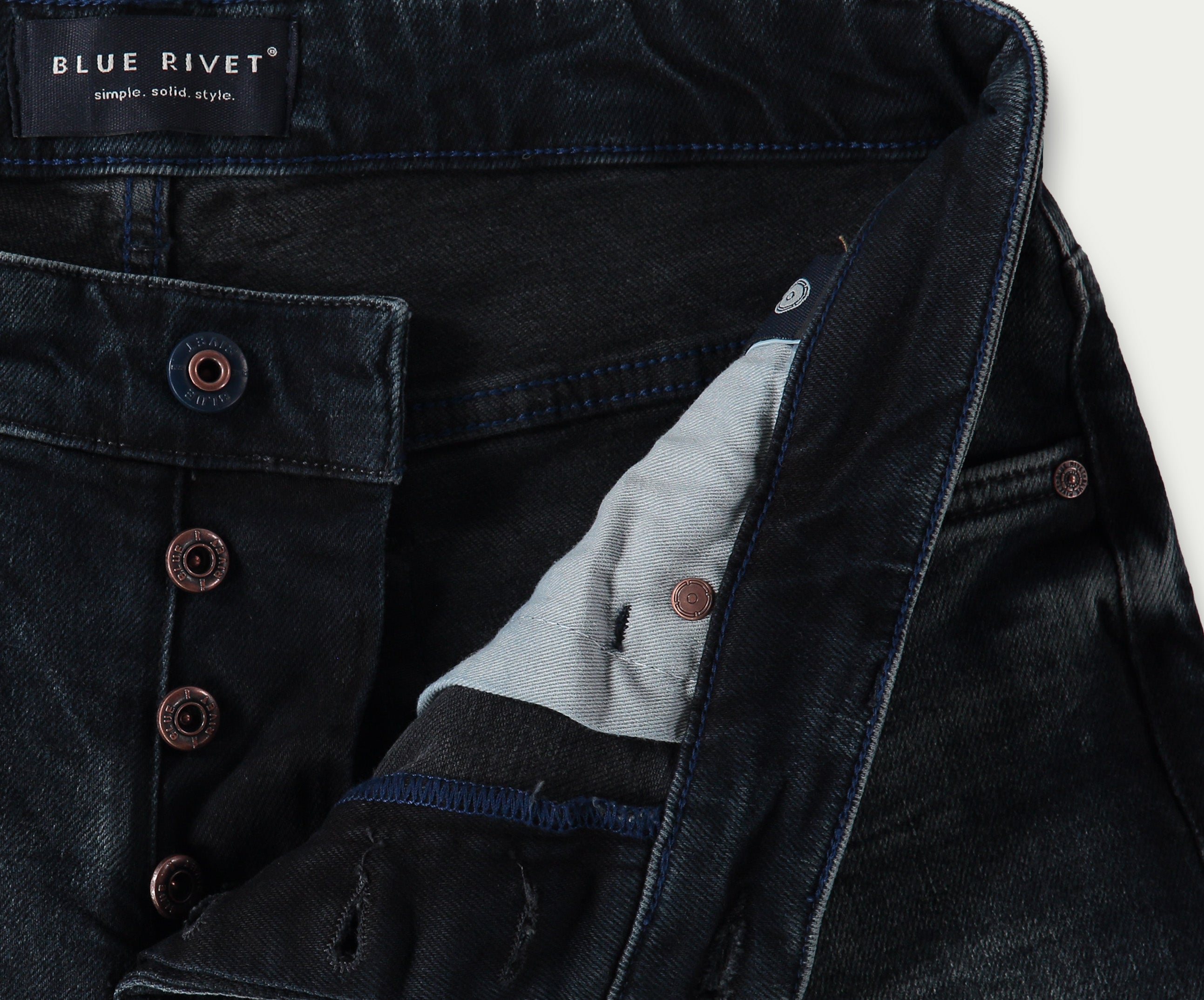 Close-up van donkerblauwe slim jeans met een zichtbare knoopsluiting. Het label luidt Blue Rivet Jeans met de tagline simple. solid. style. Koperen klinknagelknopen zijn prominent aanwezig en de binnenstof heeft een contrasterende lichtere kleur. De N01 Slim Blue Black Used spijkerbroeken bieden precies de juiste stretch voor comfort en pasvorm.
