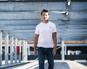 Een man in een regular fit, marineblauwe The N01 Tee van Blue Rivet Jeans en jeans stapt zelfverzekerd door een parkeergarage. Metalen balken en een hek van gaas zorgen voor een stedelijke, industriële achtergrond.