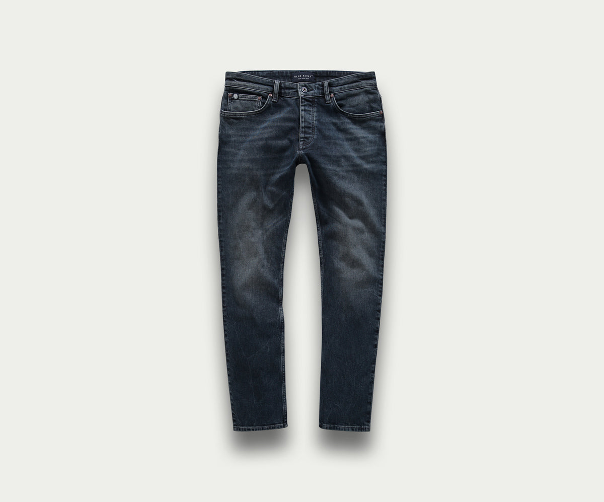 Introductie van de N01 Slim Blue Black van Blue Rivet Jeans: een premium paar slim-fit jeans in donkerblauw, met een subtiele fade op de dijen. Deze jeans zijn gemaakt met klassieke stiksels, vijf zakken en een knoopsluiting, elegant gepresenteerd tegen een lichtgrijze achtergrond.