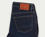 Een paar The N01 Slim Dark Indigo jeans van Blue Rivet Jeans wordt gevouwen op een lichte achtergrond getoond. Het ontwerp heeft een klein label op de achterzak en een bruine leren patch met het Blue Rivet-logo op de tailleband, wat zowel klassieke stijl als comfortabele stretch biedt.