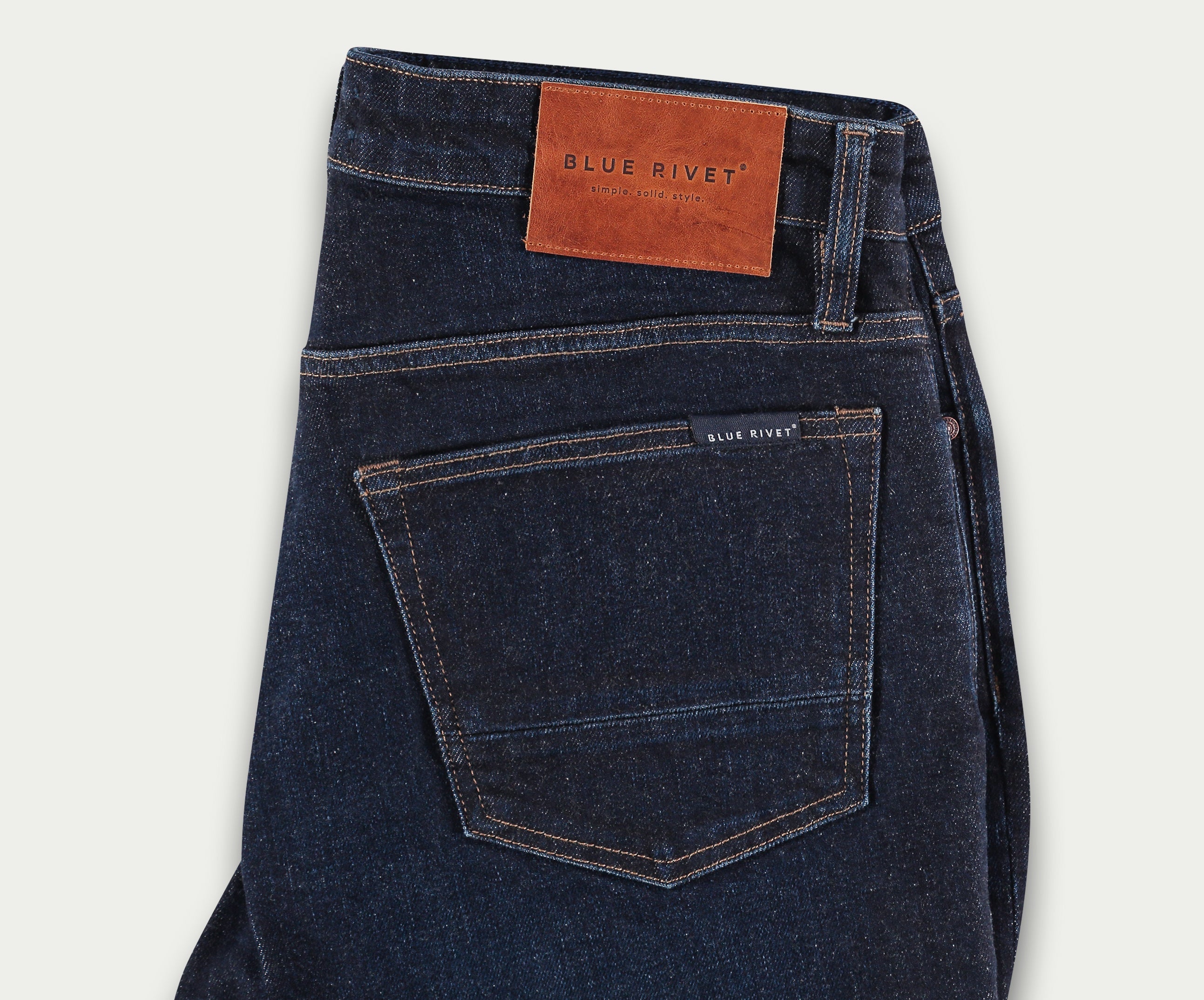 Een paar The N01 Slim Dark Indigo jeans van Blue Rivet Jeans wordt gevouwen op een lichte achtergrond getoond. Het ontwerp heeft een klein label op de achterzak en een bruine leren patch met het Blue Rivet-logo op de tailleband, wat zowel klassieke stijl als comfortabele stretch biedt.