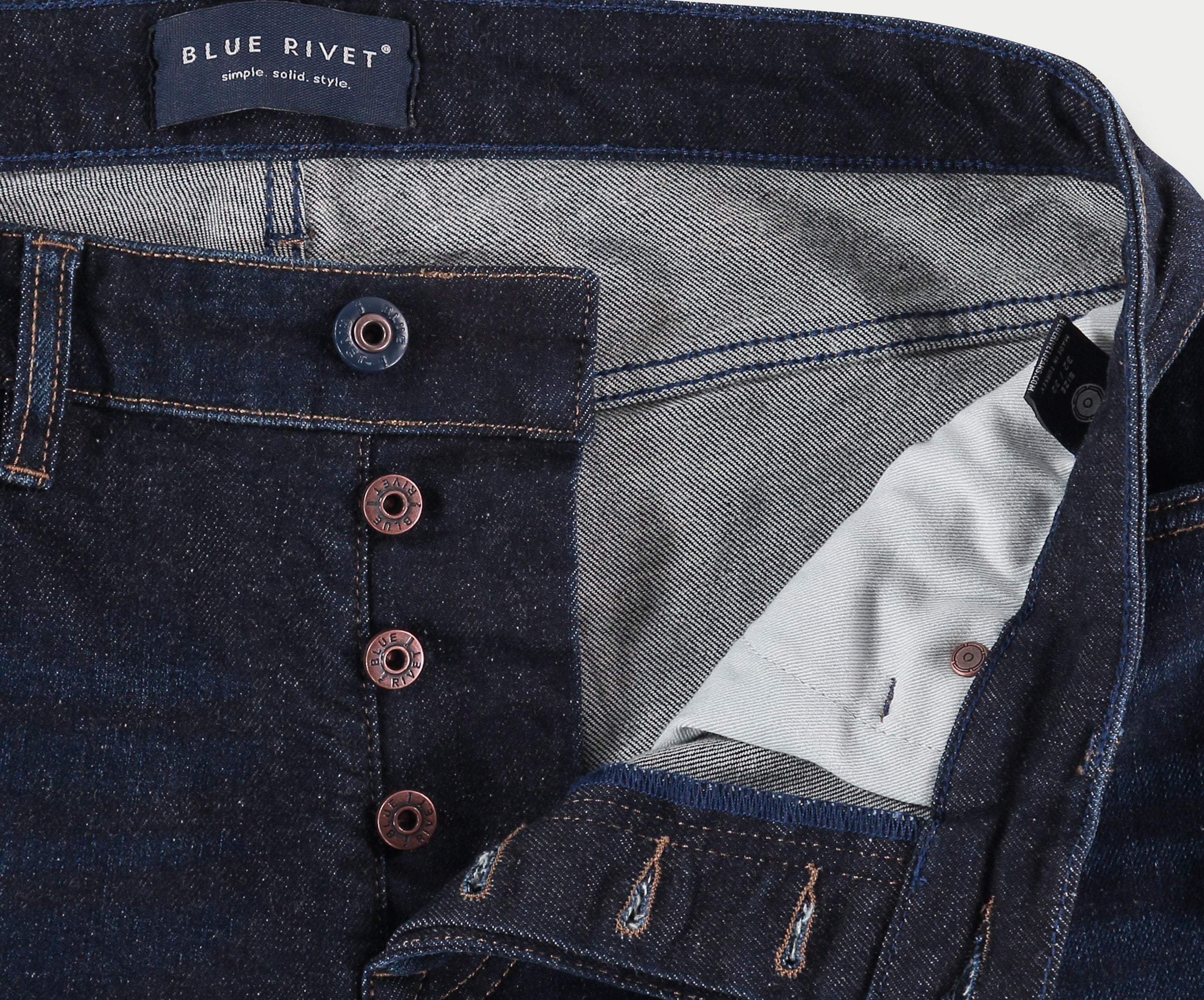 Close-up van de N01 Slim Dark Indigo jeans van Blue Rivet Jeans, met een knoopsluiting. De spijkerbroek is gedeeltelijk open om een lichtere contrasterende stoffen voering en een label aan de binnenkant van de tailleband te onthullen waarop Blue Rivet Jeans staat. Zichtbare stiksels omringen de knopen en zakken.