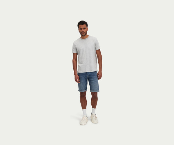 Een man staat tegen een effen witte achtergrond en draagt een lichtgrijs T-shirt, The N01 Short Dark Indigo Vintage van Blue Rivet Jeans, witte sokken en witte sneakers. Hij heeft krullend haar en een ontspannen houding.
