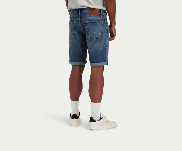 Een persoon in een grijs T-shirt en witte sneakers staat met zijn rug naar de camera en draagt de N01 Short Dark Indigo Vintage van Blue Rivet Jeans, met een omgeslagen zoom en een zichtbare bruine leren patch op de tailleband, aangevuld met witte sokken.