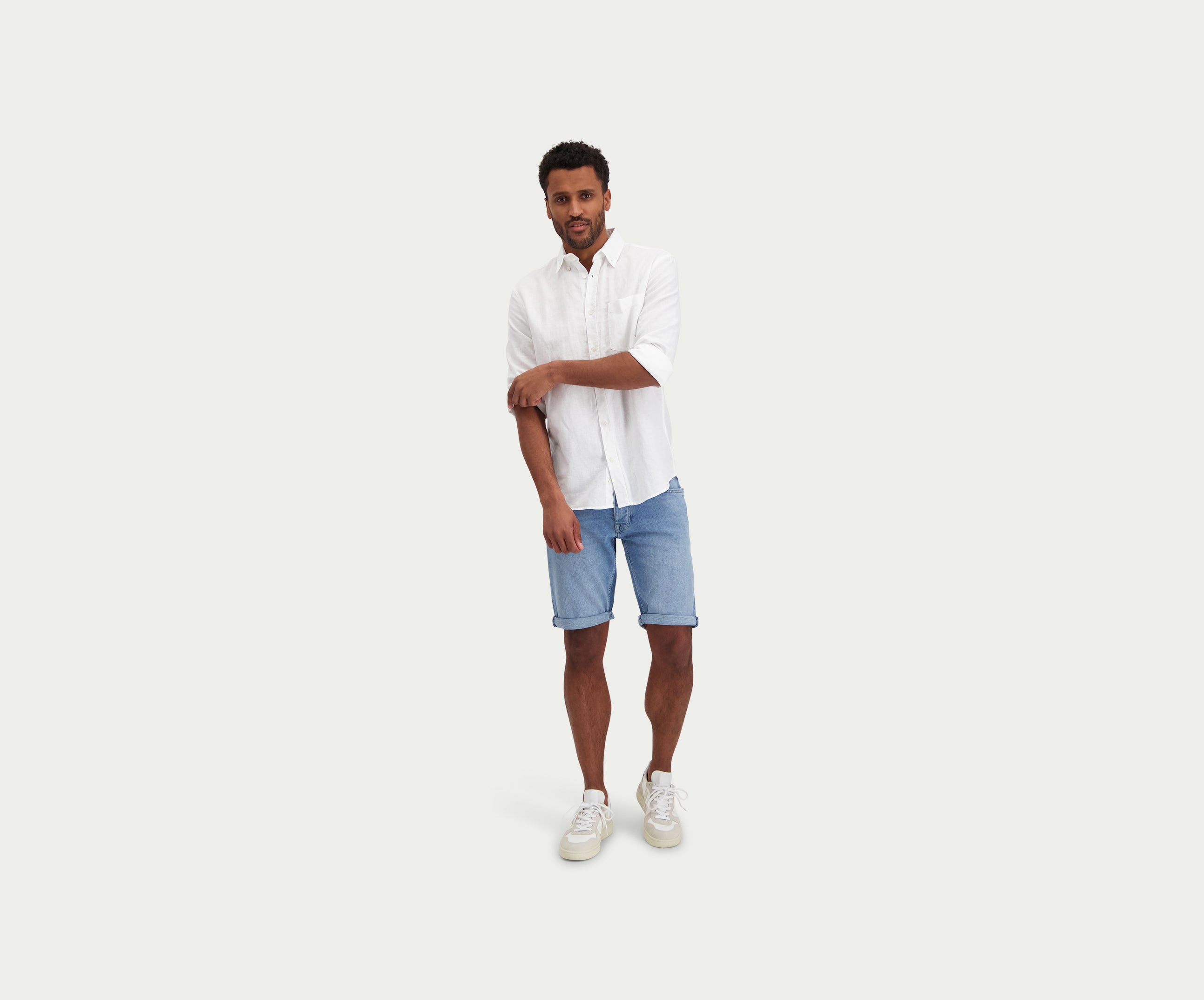 Een man met een wit overhemd en The N01 Short Stone Bleached by Blue Rivet Jeans staat tegen een effen achtergrond. Hij heeft één mouw gedeeltelijk opgerold en combineert zijn outfit met witte sneakers.