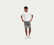 Een man met een wit poloshirt, The N01 Short Light Grey Used by Blue Rivet Jeans, en witte sneakers staat tegen een effen witte achtergrond. Hij kijkt naar voren met zijn handen op zijn heupen en kijkt lichtjes opzij.