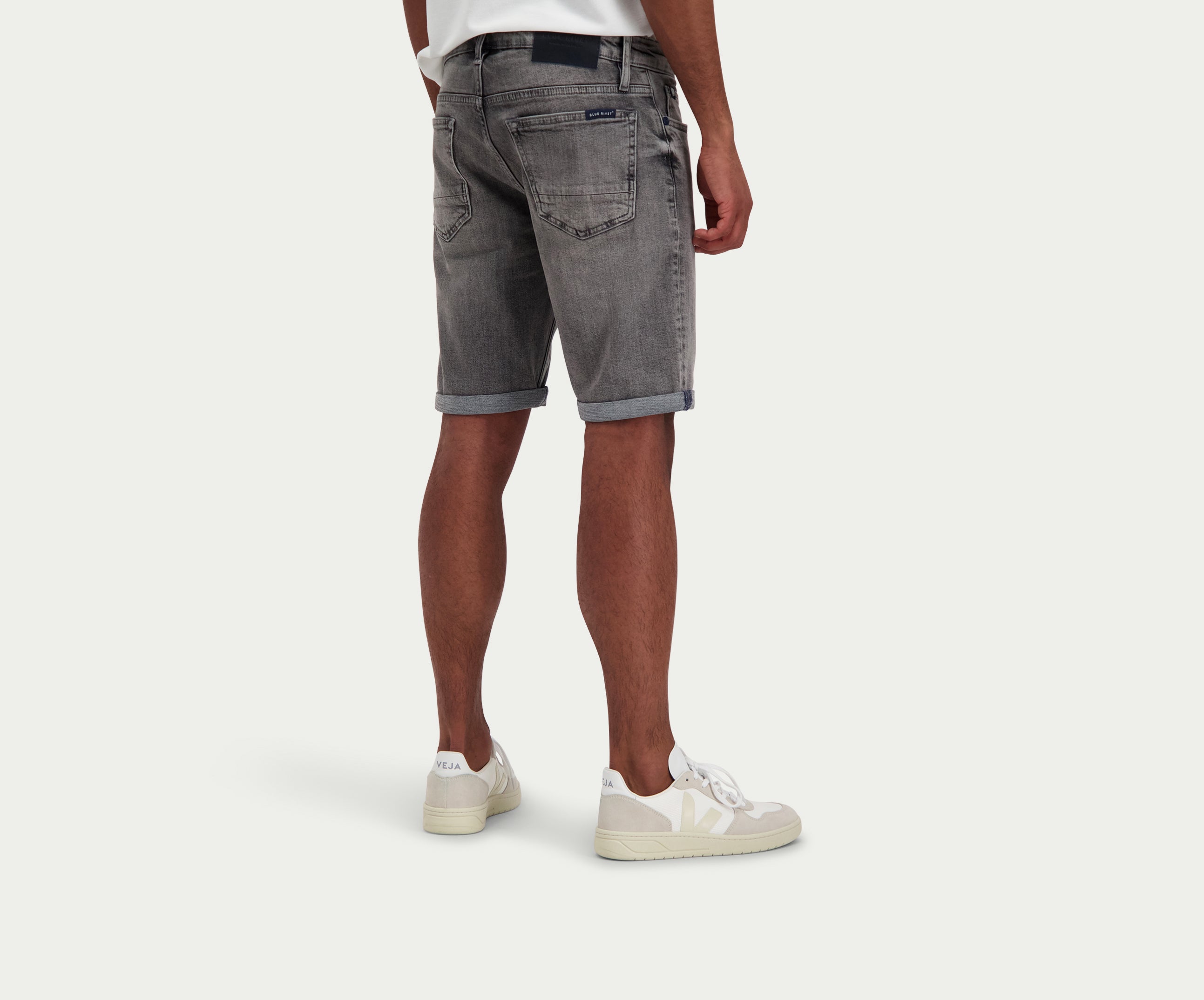 Een persoon die een wit t-shirt draagt, The N01 Short Light Grey Used by Blue Rivet Jeans in grijze denim, en off-white sneakers staat tegen een effen witte achtergrond. De afbeelding richt zich op de achterkant van de outfit, waarbij de opgerolde zoom van de shorts wordt benadrukt.