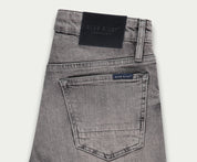 Een paar lichtgrijze jeans netjes opgevouwen, met een achterzak met een klein label met de tekst Blue Rivet Jeans. Een zwarte leren patch siert de tailleband, wat een vleugje elegantie toevoegt aan deze veelzijdige denim essentials die bekend staan als The N01 Short Light Grey Used.