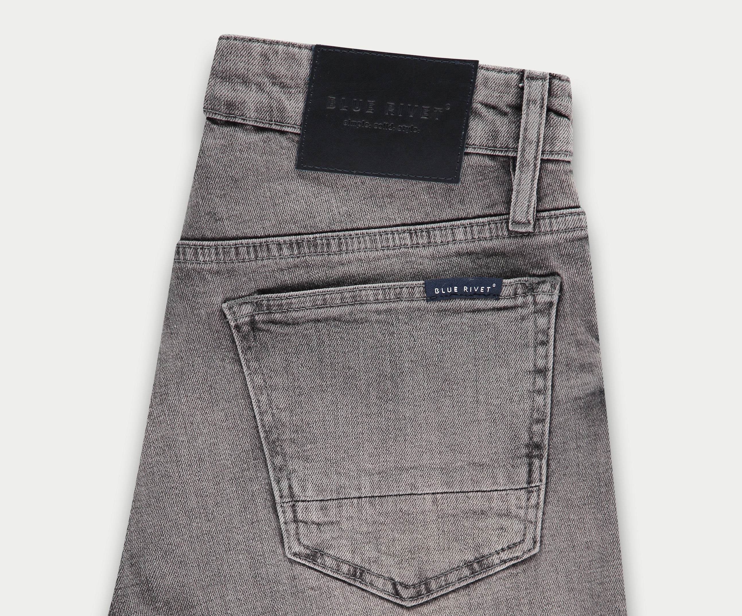 Een paar lichtgrijze jeans netjes opgevouwen, met een achterzak met een klein label met de tekst Blue Rivet Jeans. Een zwarte leren patch siert de tailleband, wat een vleugje elegantie toevoegt aan deze veelzijdige denim essentials die bekend staan als The N01 Short Light Grey Used.